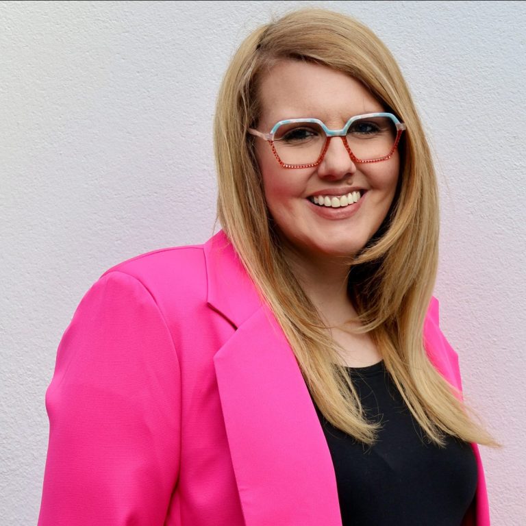 Frau mit langen blonden Haaren, rosa Blazer und Brille, lächelt vor hellen Hintergrund.